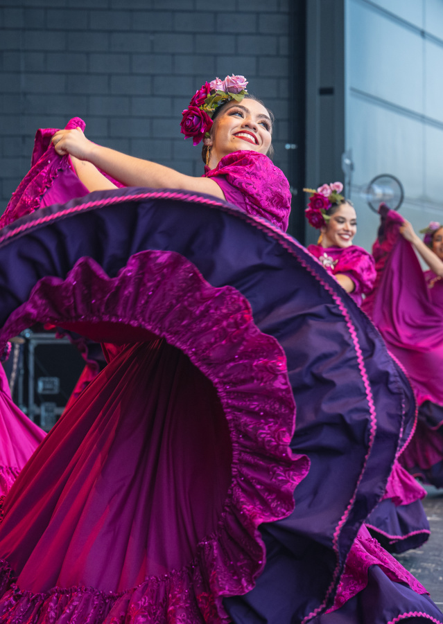 A group of female dancers performs a traditional Mexican folklórico dance on stage. They wear vibrant magenta dresses with purple ruffles and floral headpieces. The lead dancer in the foreground smiles brightly while gracefully twirling her flowing skirt. The dancers behind her mirror the movement, creating a colorful, energetic scene against a modern stage backdrop.