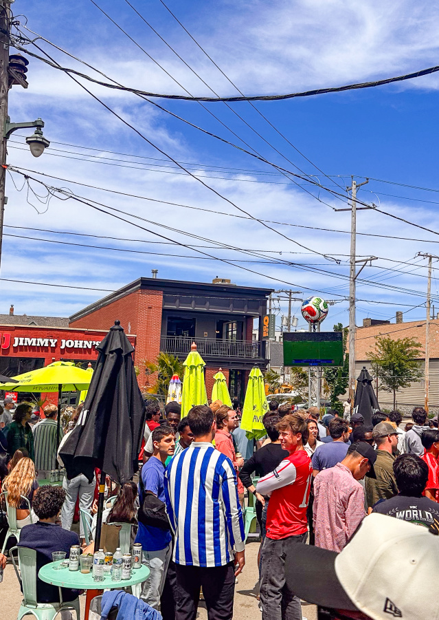 Soccer fans pack the patio and surrounding area outside Nomad World Pub on Brady Street in Milwaukee, watching a match on a large outdoor screen during a sunny afternoon.