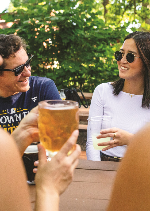 Friends gather around a picnic table at Hubbard Park Beer Garden, chatting and holding drinks beneath leafy trees on a sunny afternoon. Several people wear sunglasses while laughing and talking in the shaded outdoor seating area.