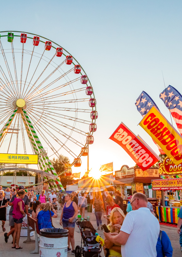 A colorful summer fair scene at sunset featuring a large Ferris wheel with green, yellow, red, and orange gondolas in the background. The foreground is bustling with people walking, eating, and enjoying the fair. Brightly lit food stands advertise corn dogs, lemonade, and "Super Dogs," with American flags waving above. A yellow sign at the Ferris wheel entrance reads "$6 PER PERSON." The sun sets behind the attractions, casting a warm glow over the festive atmosphere.