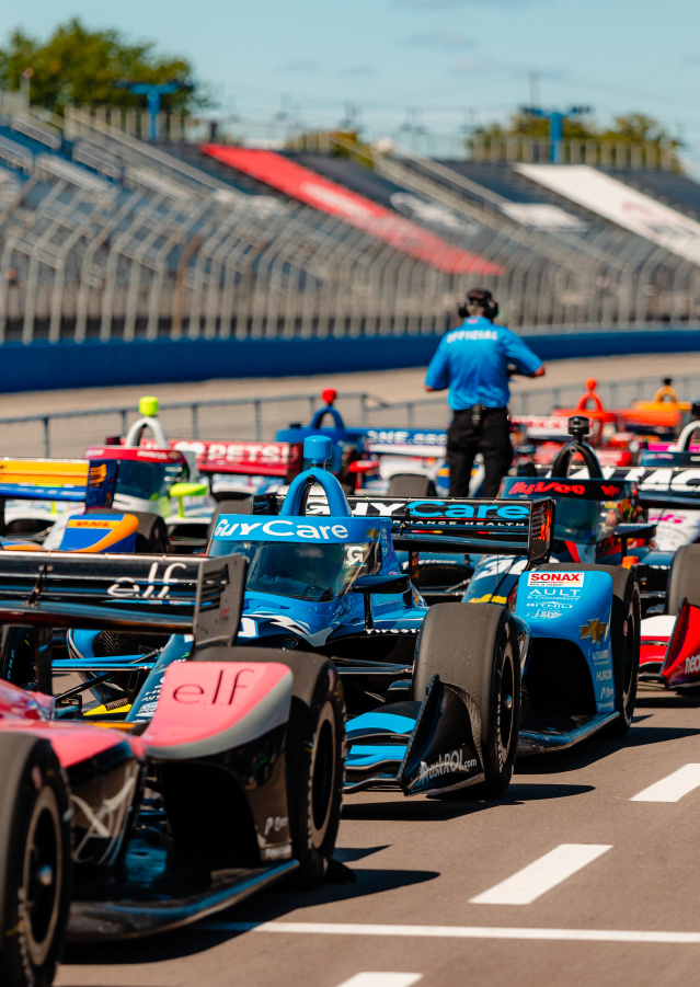 IndyCar race cars lined up on the track at the Milwaukee Mile with drivers and officials preparing for the start of the race, as spectators watch from the grandstands under a sunny sky.