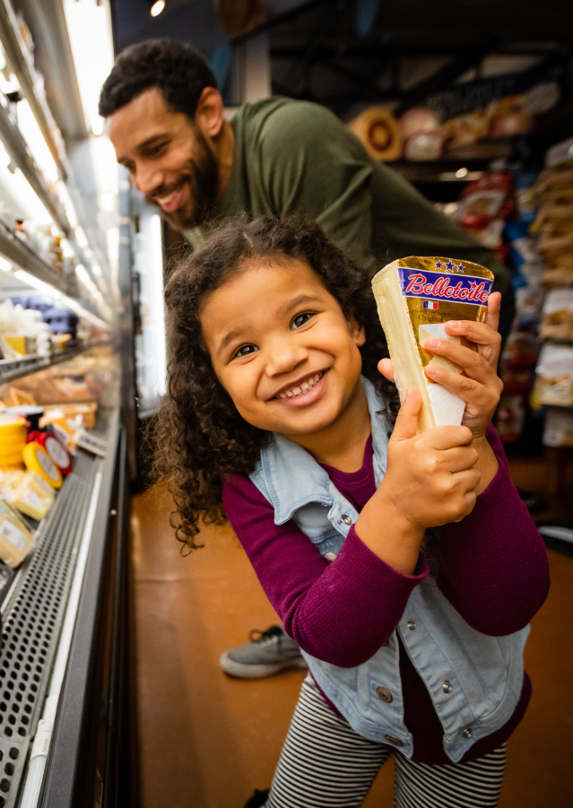 a little girl showing off a wedge of cheese in front of a large deli cooler