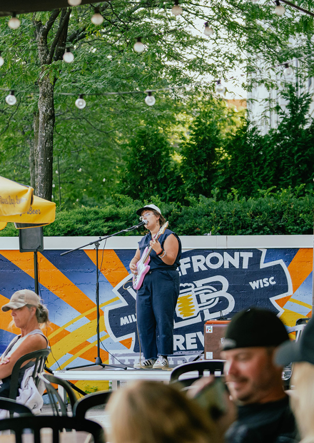 A woman in a navy blue romper singing into a microphone on a stage, holding a pink guitar, with people in front of her sitting at tables under gold and blue umbrellas on a patio along the riverfront with trees in the background.