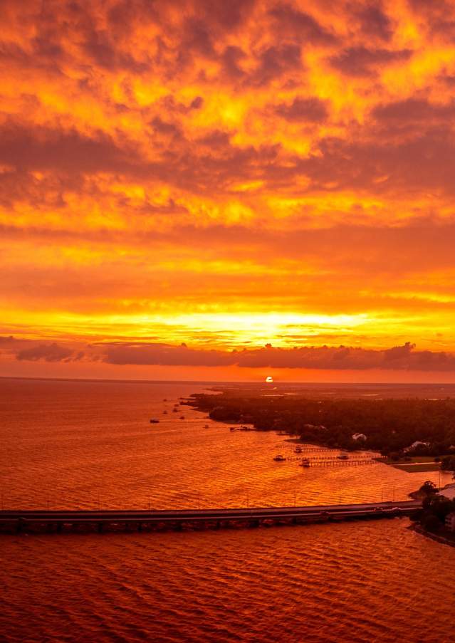 Aerial view of Lake Pontchartrain and the Mandeville lakefront beneath a vivid orange and gold sunset, with the Causeway stretching across the water.