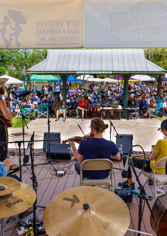 A band performs on the Abita Trailhead Museum stage beneath a pavilion while a seated crowd watches during the Abita Springs Busker Festival.