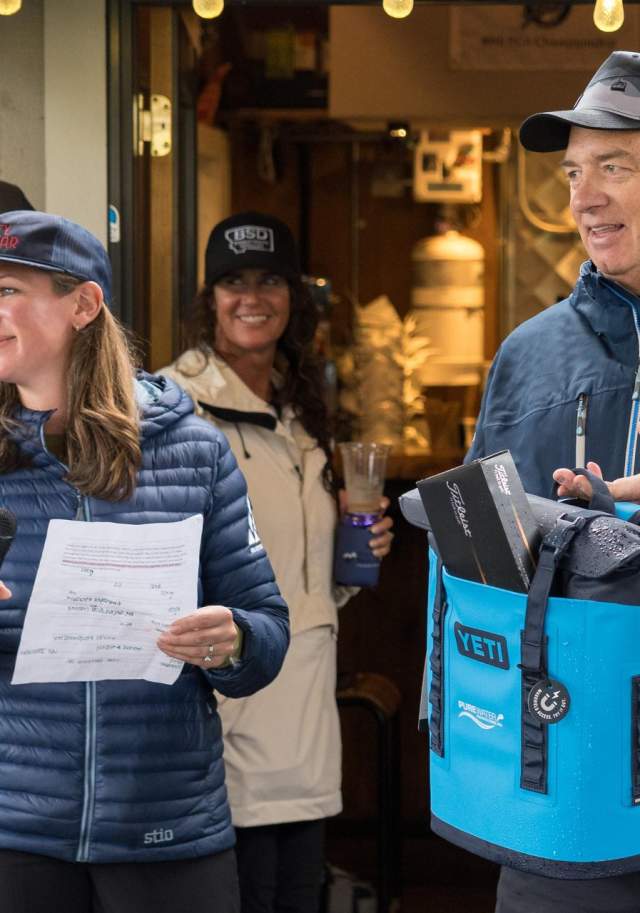 A woman with a microphone and a man holding a blue bag stand in front of a cozy venue, engaging an audience. Others are visible in the background.