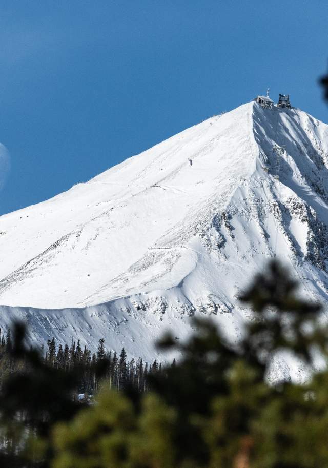 Snow-covered Lone Mountain and the moon over a dark evergreen forest in Big Sky, Montana.