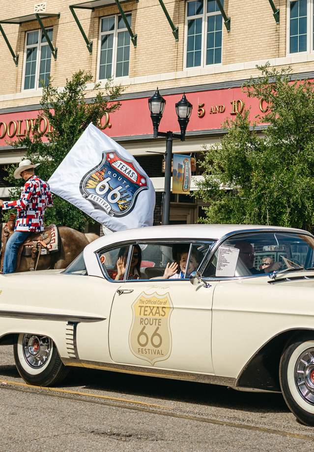 cream colored classic cadillac with a Route 66 shield on the side of it. The car is called the yellow rose and is the offical vehicle of the tezas route 66 festival. Man riding a horse holdinga. flag with the festival logo in the background