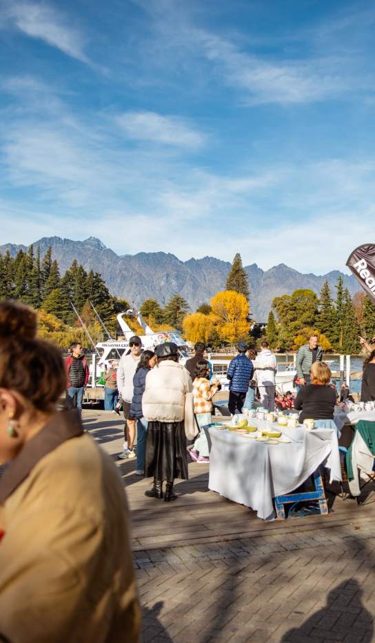 People browsing stalls at Queenstown craft market with autumnal trees and the Remarkables mountain range in the background