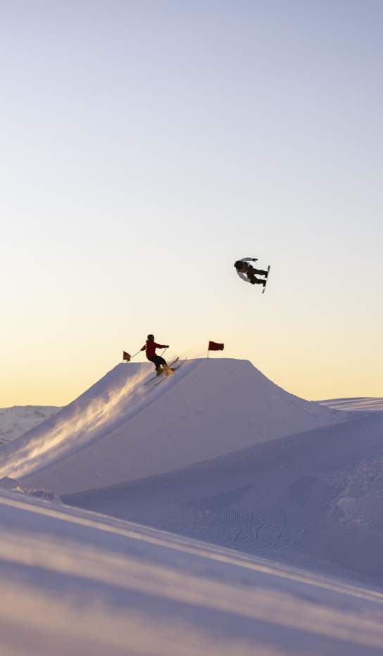 Snowboarders and a skier perform aerial tricks off a jump at Cardrona Alpine Resort at sunrise.