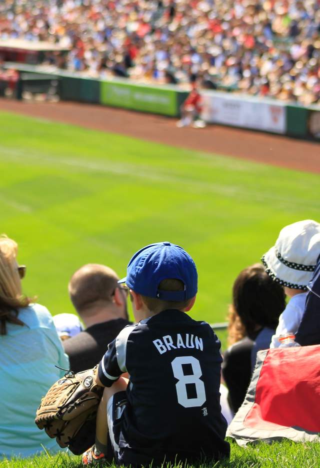 Kid sitting in a crowd at a baseball game with jersey and glove on.