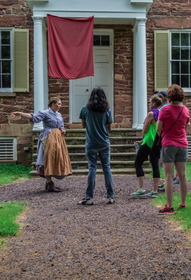 A group of visitors listening to a woman dressed in historic attire outside of a brick building