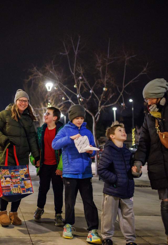 A group of two adults and three children walk together on a decorated city sidewalk at night during the holiday season. They are bundled in winter coats, hats, and gloves. Festive lights and small decorated trees line the shop windows on the left. One adult carries a colorful tote bag, and one child holds a snack. Everyone appears cheerful and engaged with one another as they enjoy the winter evening.”