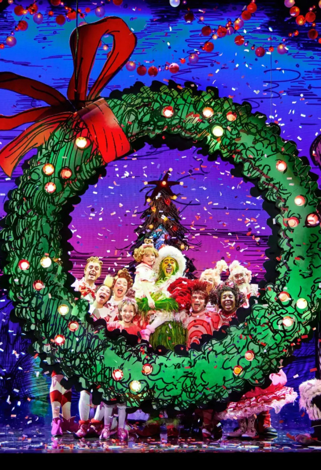 A colorful stage scene from a Christmas-themed musical featuring characters in bright costumes gathered around a giant green wreath with a red bow. Snow-like confetti falls from above, and whimsical, snow-covered houses are visible in the background.