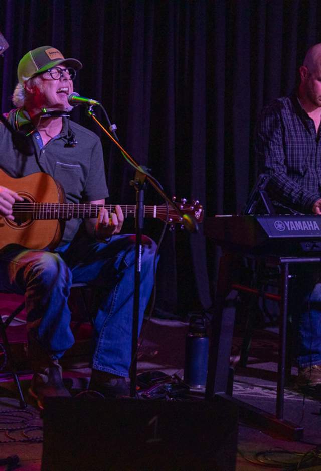man wearing a ballcap playing a guitar and singing, and another man playing keyboards