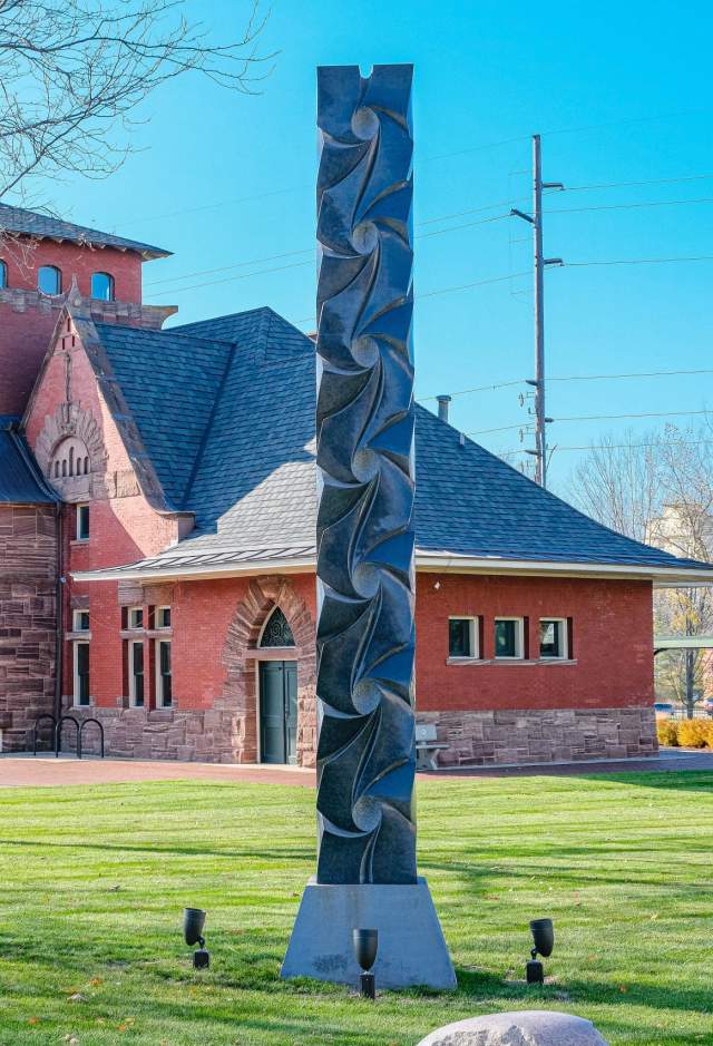 Black granite column sculpture "all my relations" sits on lawn in front of historic brick building.
