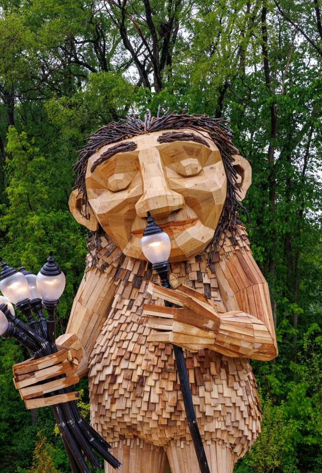 Large Wooden Troll sculpture smaelling lamps as if they were flowers against green trees.