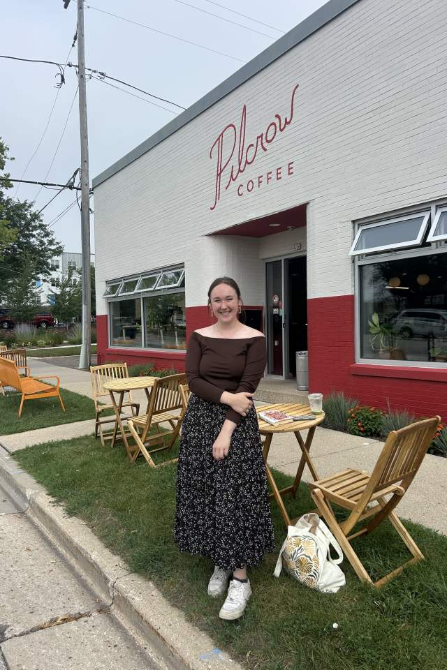 A woman smiles while standing outside Pilcrow Coffee, a café with outdoor seating including wooden tables and chairs set up along the sidewalk.