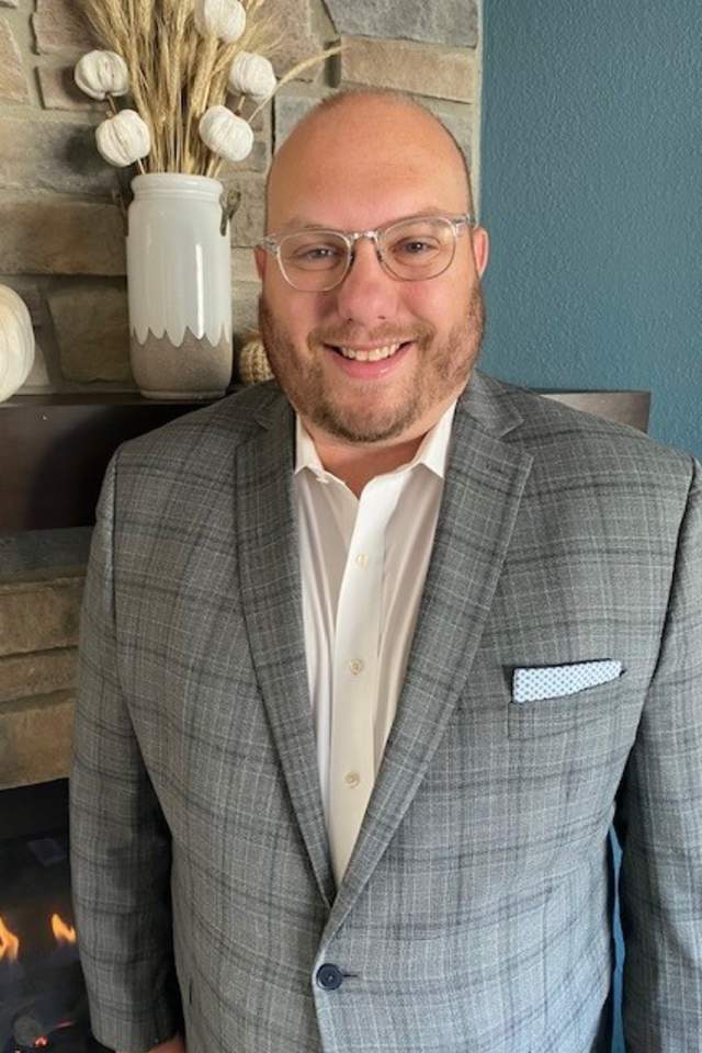 Ben Risseeuw in glasses smiles at the camera, wearing a gray plaid blazer over a white shirt, standing indoors near a stone wall with neutral décor.