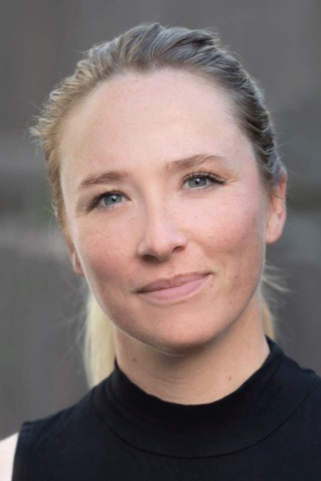 Portrait of a woman with blonde hair tied back, wearing a sleeveless black top, looking at the camera with a neutral expression. The background is softly blurred.