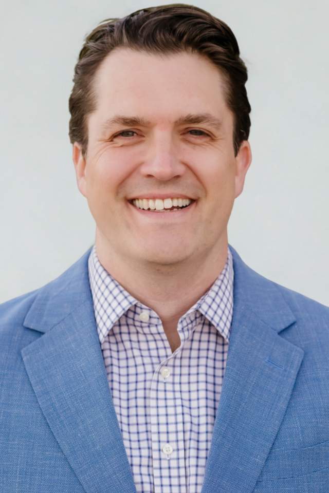 smiles against a clean, neutral background, wearing a light blue blazer over a patterned button-down shirt for a polished, professional portrait.