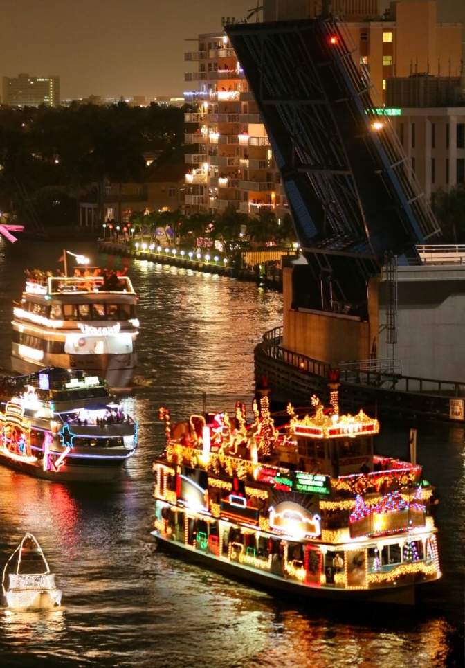 Several Boats of different sizes all lit up and decorated for the December holidays floating up the river through an open draw-bridge