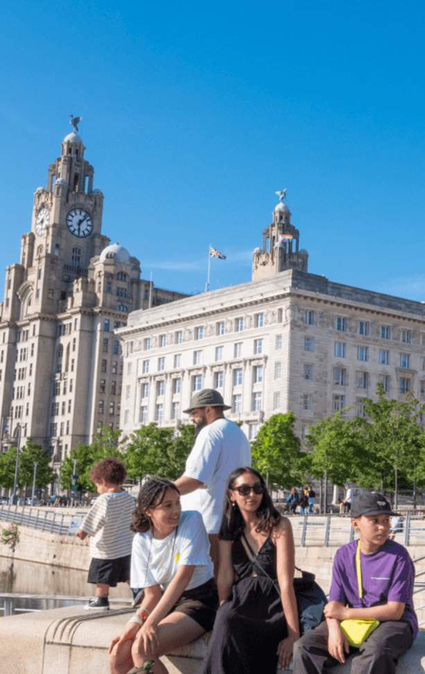 Liverpool Pier Head in the sunshine with a family sitting on a wall. A small child is holding a balloon in the shape of a rainbow.