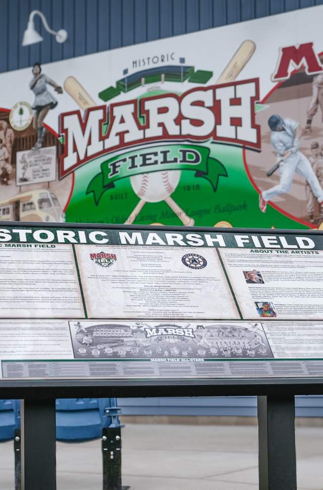 Informational display at Historic Marsh Field with detailed panels about the baseball field's history, set against a mural featuring baseball imagery and logos.