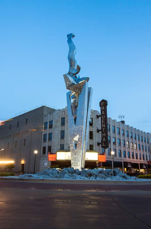 steel sculpture "muskegon rising" sits in roundabout in front of historic Frauenthal theater at sunset. blurred car headlights illuminate the left side of the photo
