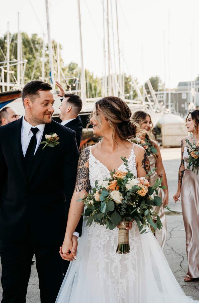 young groom and bride hold hand while looking at one another. int the background are groomsmen, bridesmaids and boats in a marina