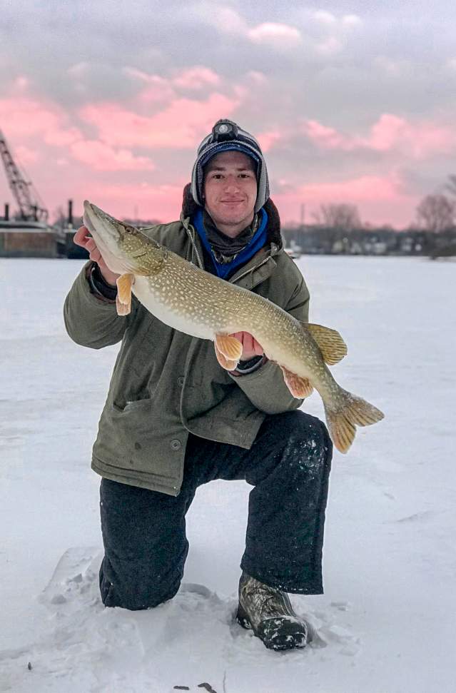 A person in winter attire holding a large Northern Pike while kneeling on a frozen Muskegon Lake.