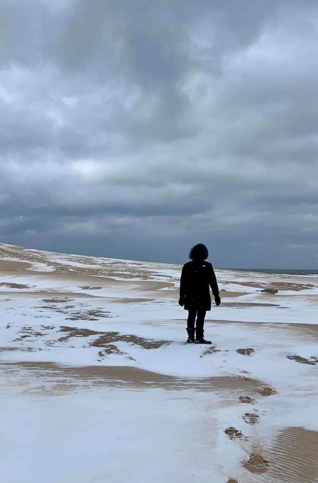 Person standing on a snow-covered beach at Muskegon State Park looking at sand dunes under a cloudy sky.