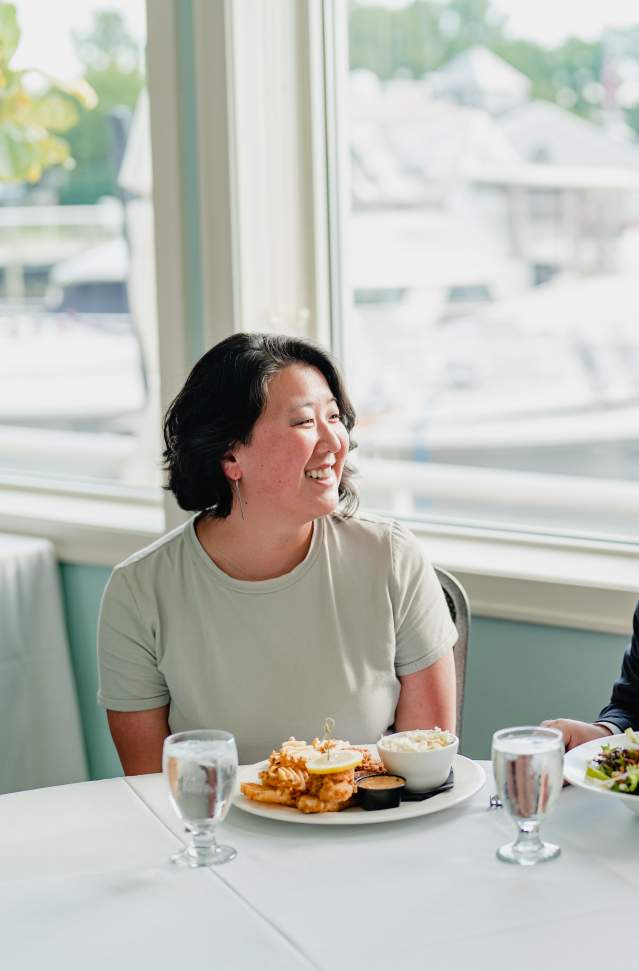 dark haired woman and bearded man smile at one another. They are seated at restaurant with plates of food before them. Docked boats can be seen through the windows behind them