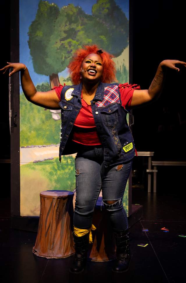 woman with orange hair acts out character on stage wearing red shirt, jean vest with colorful patches