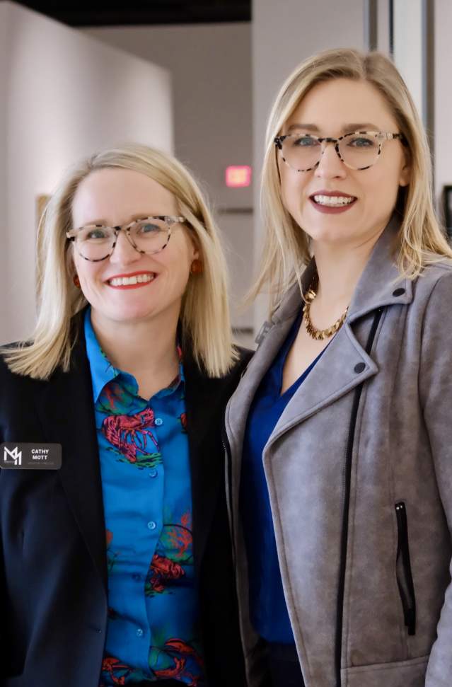 Two women smiling in a modern art gallery. Both wear glasses and business attire, exuding a professional and friendly atmosphere amidst artwork displays.