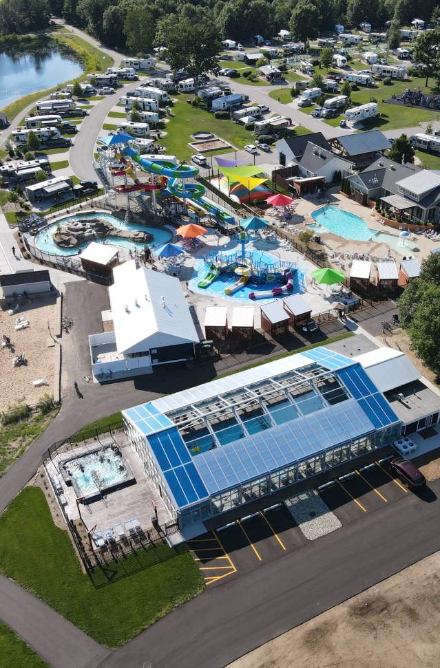 Aerial view of a bustling campsite featuring an array of colorful water slides, pools, and cabanas. Lush greenery surrounds the site near a shimmering lake, creating a lively and inviting atmosphere.