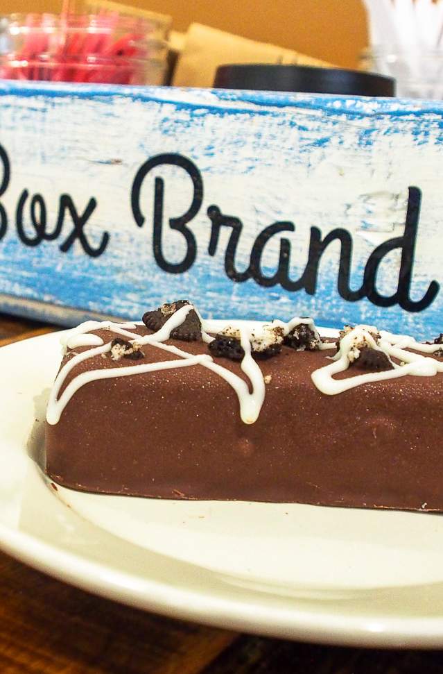 chocolate covered specialty ice cream bar sits on white plate on highly polished wooden table. in the background is a weathered blue and white box with the words ice box brand in black.