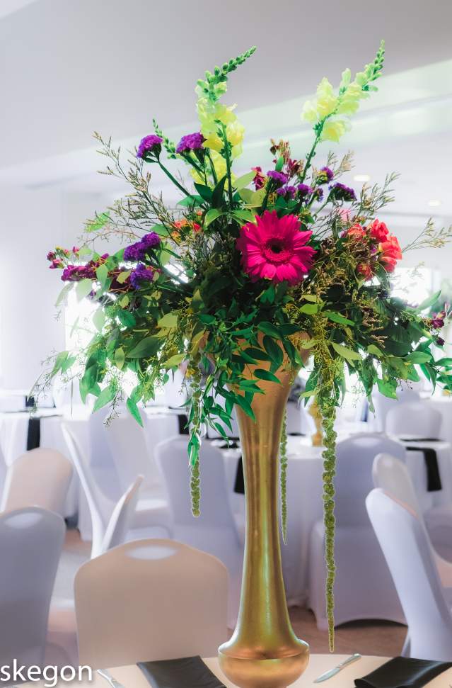 floral arrangment in tall gold vase sits event room with white table coverings and chair coverings
