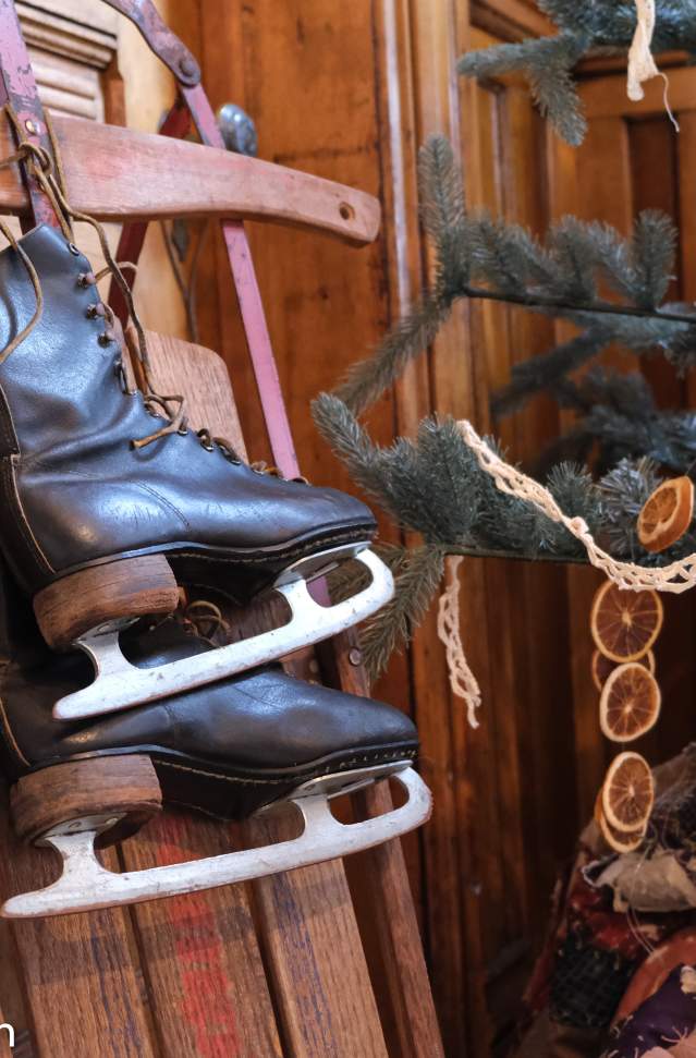 A pair of black ice skates hanging from a wooden sled, surrounded by festive decorations including a garland and dried citrus slices, inside a rustic setting.