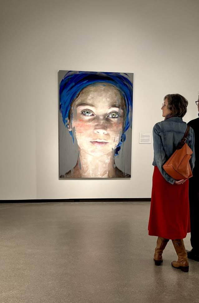 Two visitors at Muskegon Museum of Art viewing various portraits, including a prominent portrait  painting of a young woman with a blue scarf covering her head.