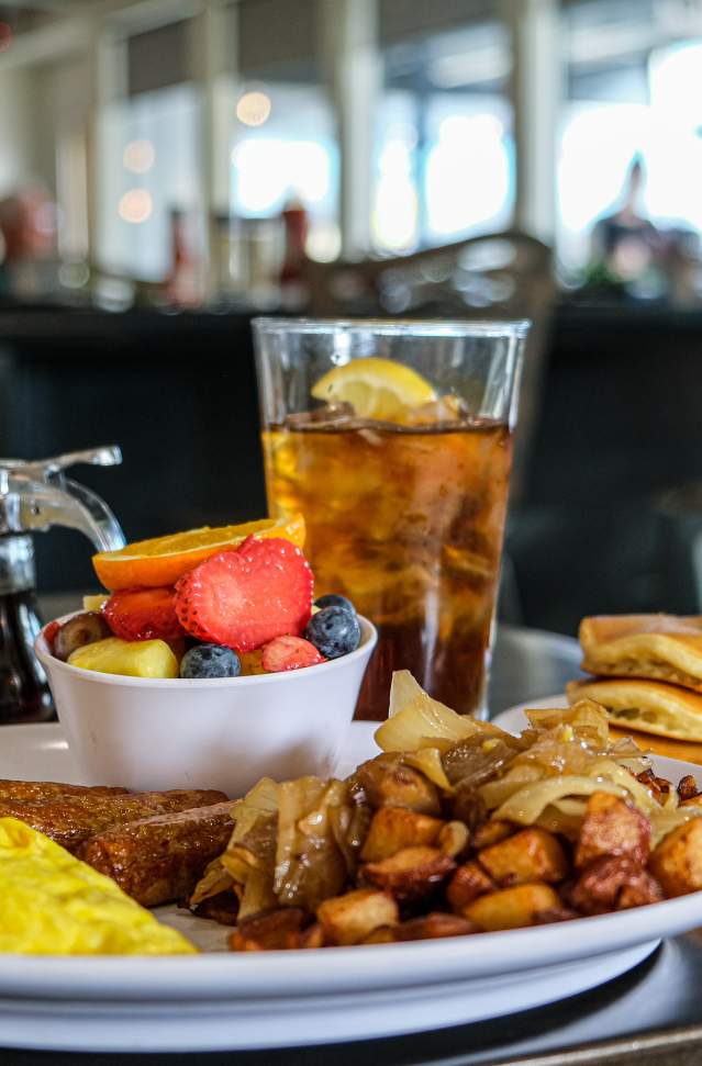 Breakfast plate with scrambled eggs, potatoes, fruit cup, sausage links. behind plate is a cup of coffee, syrup container, glass of iced tea and smallplate with stack of pancakes topped by pat of melting butter. sun filled windows are in the far background.