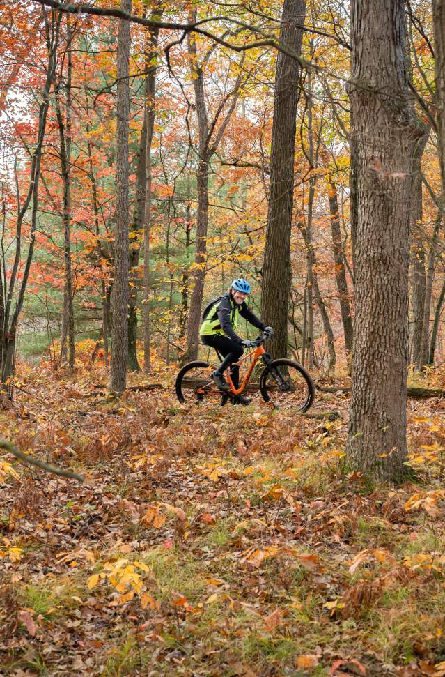 A cyclist wearing a helmet and bright clothing is riding a mountain bike on a trail amid colorful autumn trees. Fallen leaves carpet the ground.