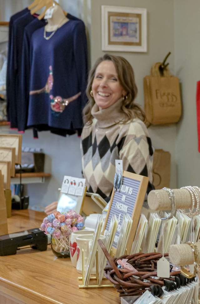 A person smiling behind the cashier counter in a boutique store called "Figgs Boutique." The store is well-stocked with various clothing items, accessories, and unique gifts displayed neatly around the shop.