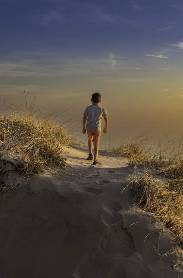 young boy ascends sand dune walking away from camera. the sky is blue fading to sunset orange and blending into the water of lake michigan.