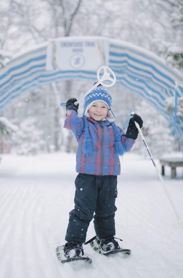 little boy in light blue stocking cap stands on snow trail. he is wearing snowshoes and holds a pole up over his head