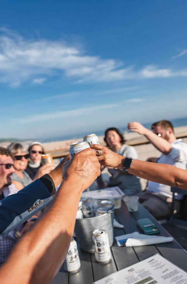 A group of people enjoying drinks and toasting together at an outdoor table with a view of the Lake Michigan under a clear blue sky.
