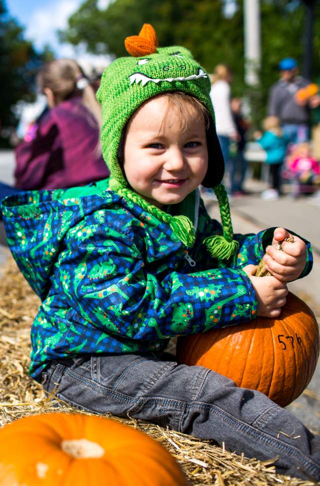 smiling child wears green knit hat and blue and green winter coat. He is holding a pumpkin and sitting on a straw bale, smiling at the camera