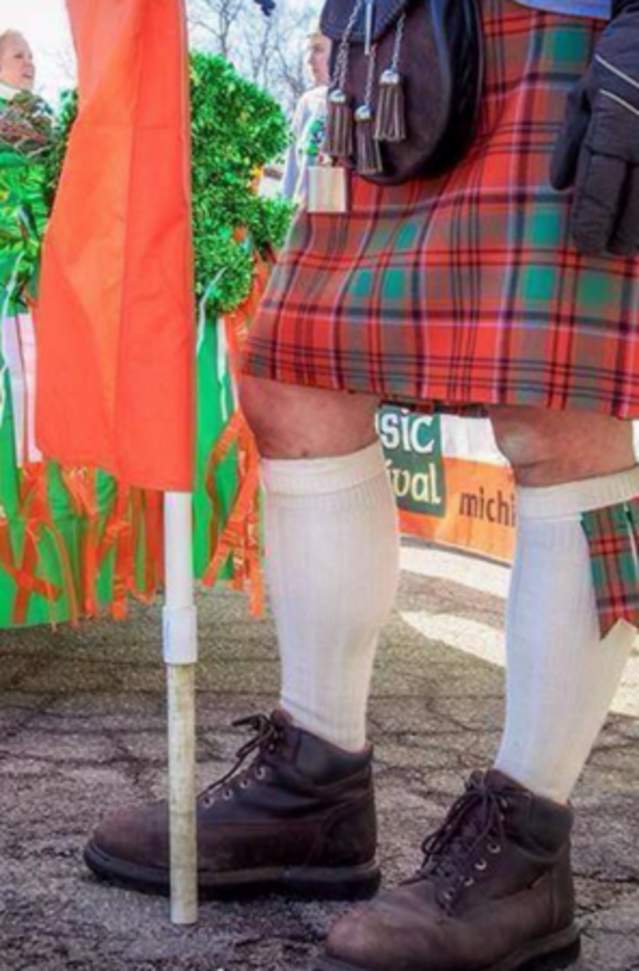 image of man from waist down holding an orange flag, wearing a traditional kilt in front of float decorated in green, white and orange with shamrocks