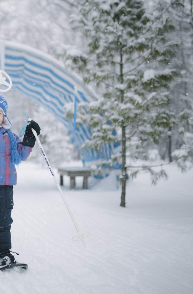 little boy in light blue stocking cap stands on snow trail. he is wearing snowshoes and holds a pole up over his head