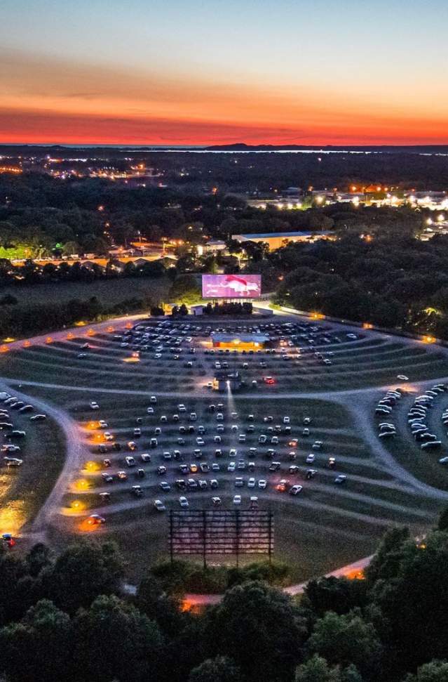 birds eye viewof Getty drive in theater at sunset in muskegon michigan shows cars parked at 4 screens surrounded by trees, city lights in distance and sunset on horizon